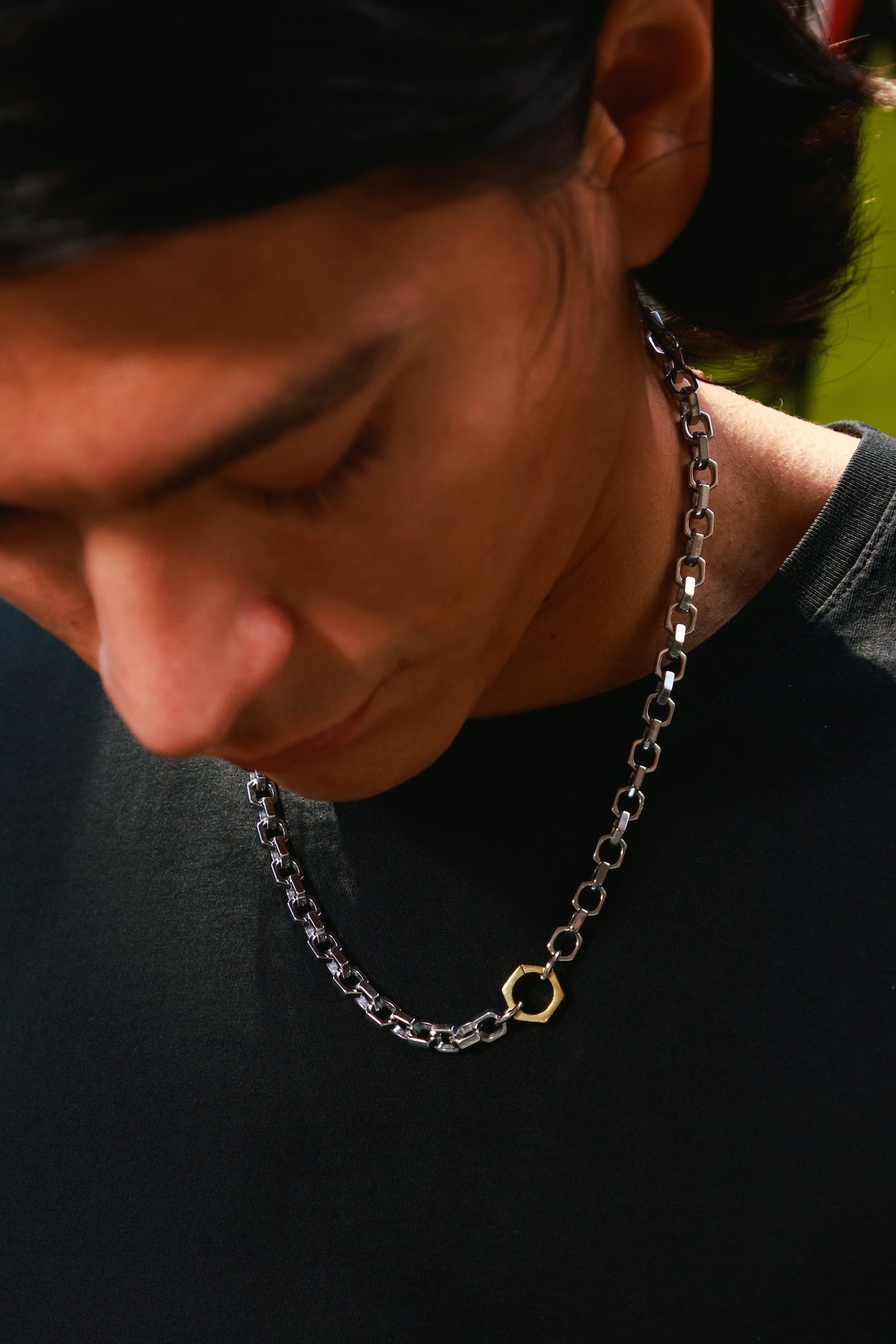 A close-up lifestyle shot of a man wearing a silver chain necklace with a small gold hexagonal pendant, looking down in warm natural light.