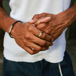 A close-up lifestyle shot of a man's hands clasped in front of his waist, wearing a white ribbed tank top, dark blue jeans, a black cord bracelet with silver hex clasp, and a gold band on his ring finger.