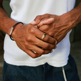 A close-up lifestyle shot of a man's hands clasped in front of his waist, wearing a white ribbed tank top, dark blue jeans, a black cord bracelet with silver hex clasp, and a gold band on his ring finger.
