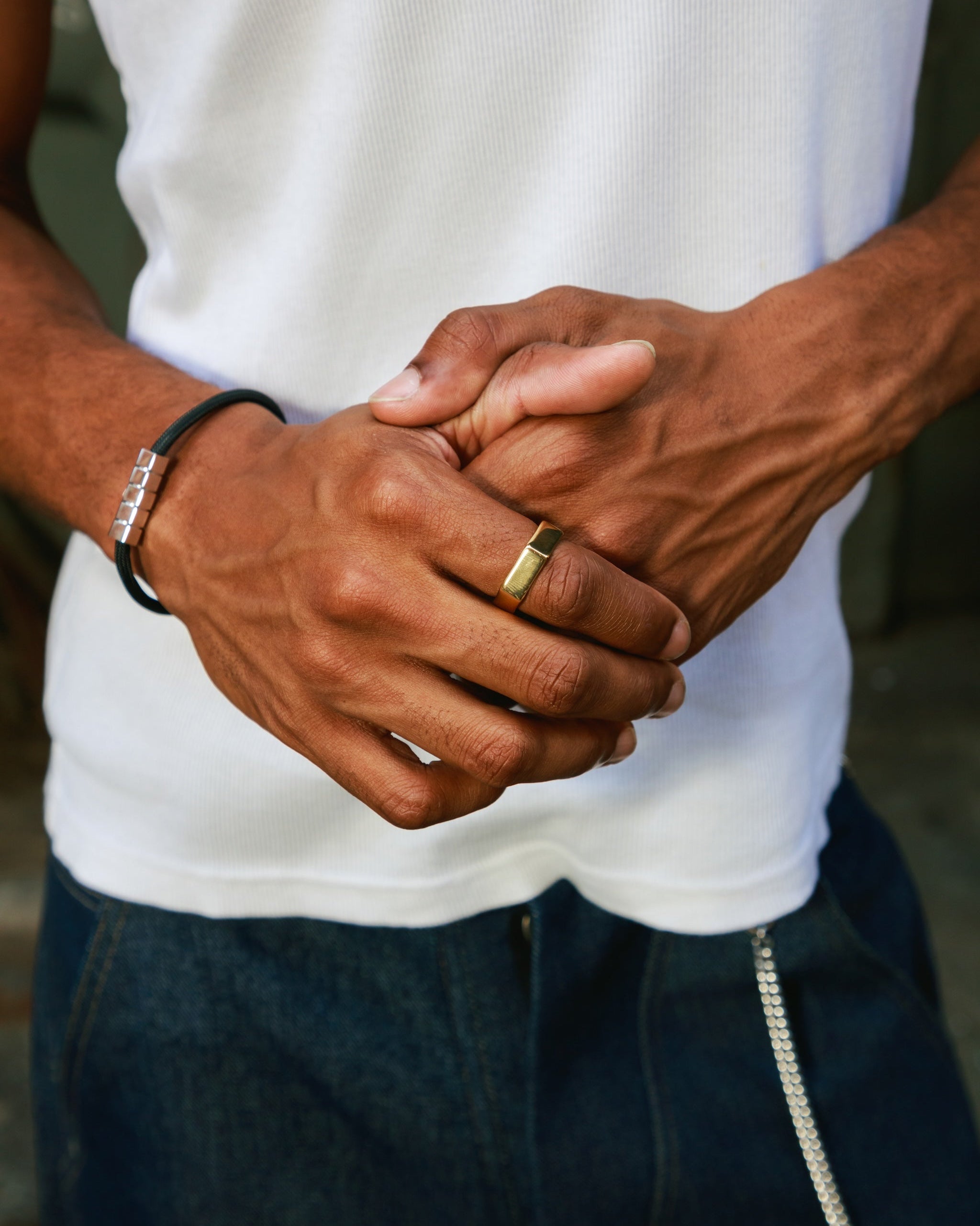 A close-up lifestyle shot of a man's hands clasped in front of his waist, wearing a white ribbed tank top, dark blue jeans, a black cord bracelet with silver hex clasp, and a gold band on his ring finger.