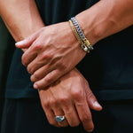 A close-up of a person's crossed arms wearing a stack of two bracelets on the right wrist, including a silver chain bracelet, a gold chain bracelet, along with a silver ring on the finger.