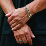 A close-up of a person's crossed arms wearing a stack of two bracelets on the right wrist, including a silver chain bracelet, a gold chain bracelet, along with a silver ring on the finger.