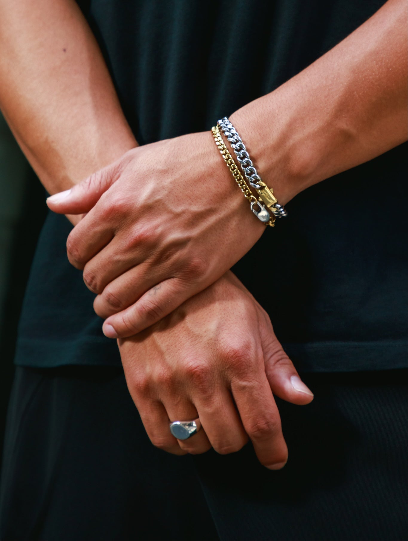 A close-up of a person's crossed arms wearing a stack of two bracelets on the right wrist, including a silver chain bracelet, a gold chain bracelet, along with a silver ring on the finger.
