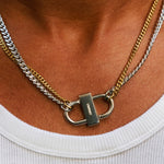 A close-up of a person's neck wearing a two-tone silver and gold chain necklace with a padlock-shaped clasp, paired with a white ribbed t-shirt.