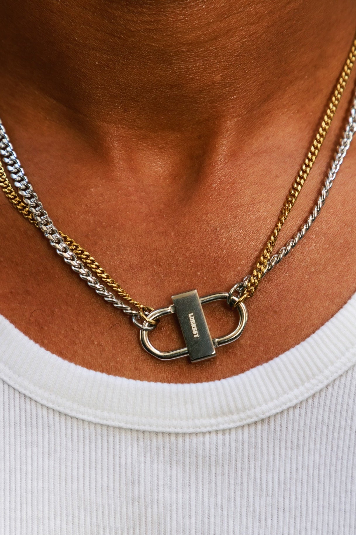 A close-up of a person's neck wearing a two-tone silver and gold chain necklace with a padlock-shaped clasp, paired with a white ribbed t-shirt.