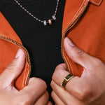 Close-up of a person wearing a silver ball chain necklace with five small hex beads (two silver, two black, one copper) over a black t-shirt and orange-brown leather jacket, with hands holding the jacket open.