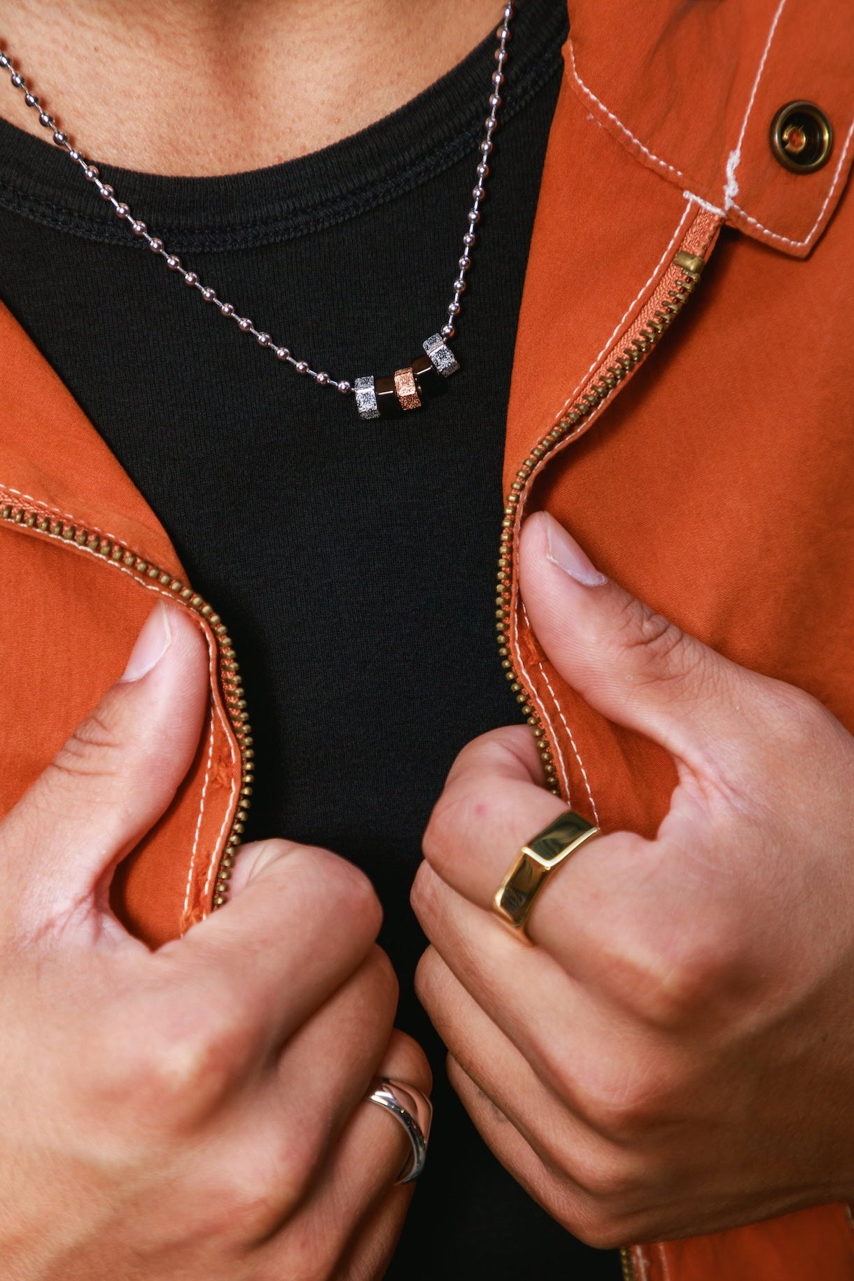 Close-up of a person wearing a silver ball chain necklace with five small hex beads (two silver, two black, one copper) over a black t-shirt and orange-brown leather jacket, with hands holding the jacket open.
