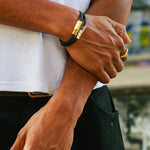 A close-up lifestyle shot of a man wearing a black leather bracelet with a gold-toned hex clasp on his wrist, paired with a white t-shirt and black pants.