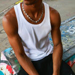 A man wearing a white ribbed tank top and black pants, sitting on a graffiti-covered bench. He is wearing a silver chain necklace and a black bracelet.