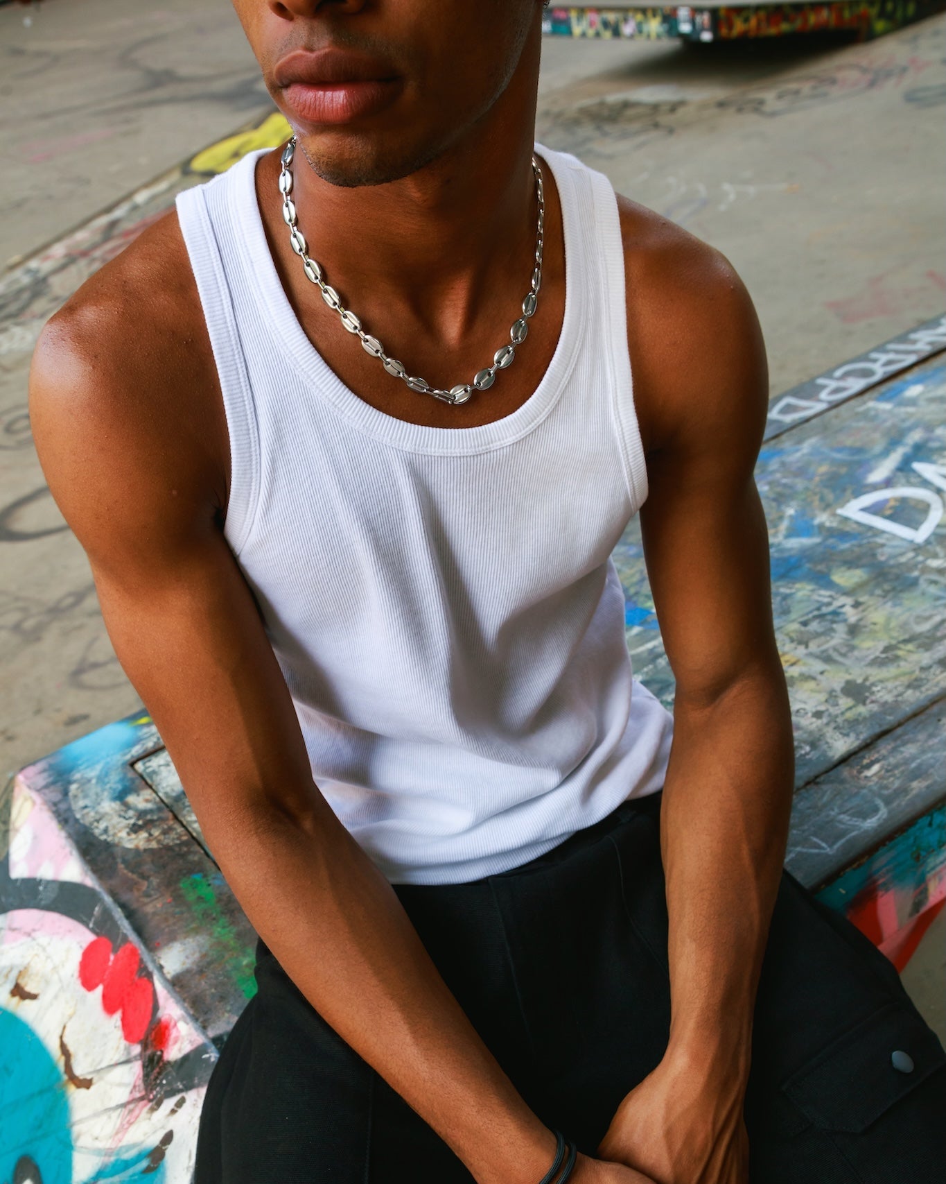A man wearing a white ribbed tank top and black pants, sitting on a graffiti-covered bench. He is wearing a silver chain necklace and a black bracelet.