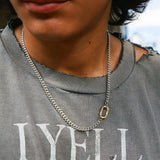 A man wearing a grey T-shirt, styled with a silver Cuban chain featuring a gold carabiner clasp.