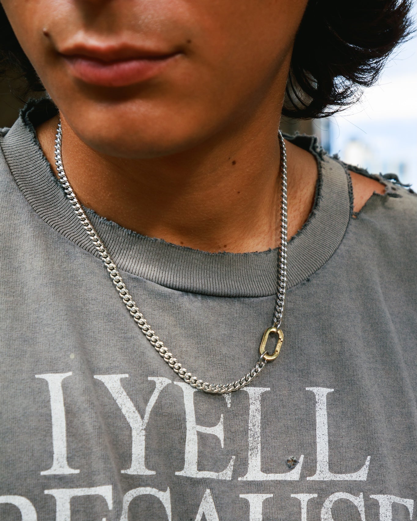 A man wearing a grey T-shirt, styled with a silver Cuban chain featuring a gold carabiner clasp.