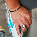 A close-up lifestyle shot of a person's hand resting on a white and teal skateboard, wearing a silver signet ring and a black cord bracelet.