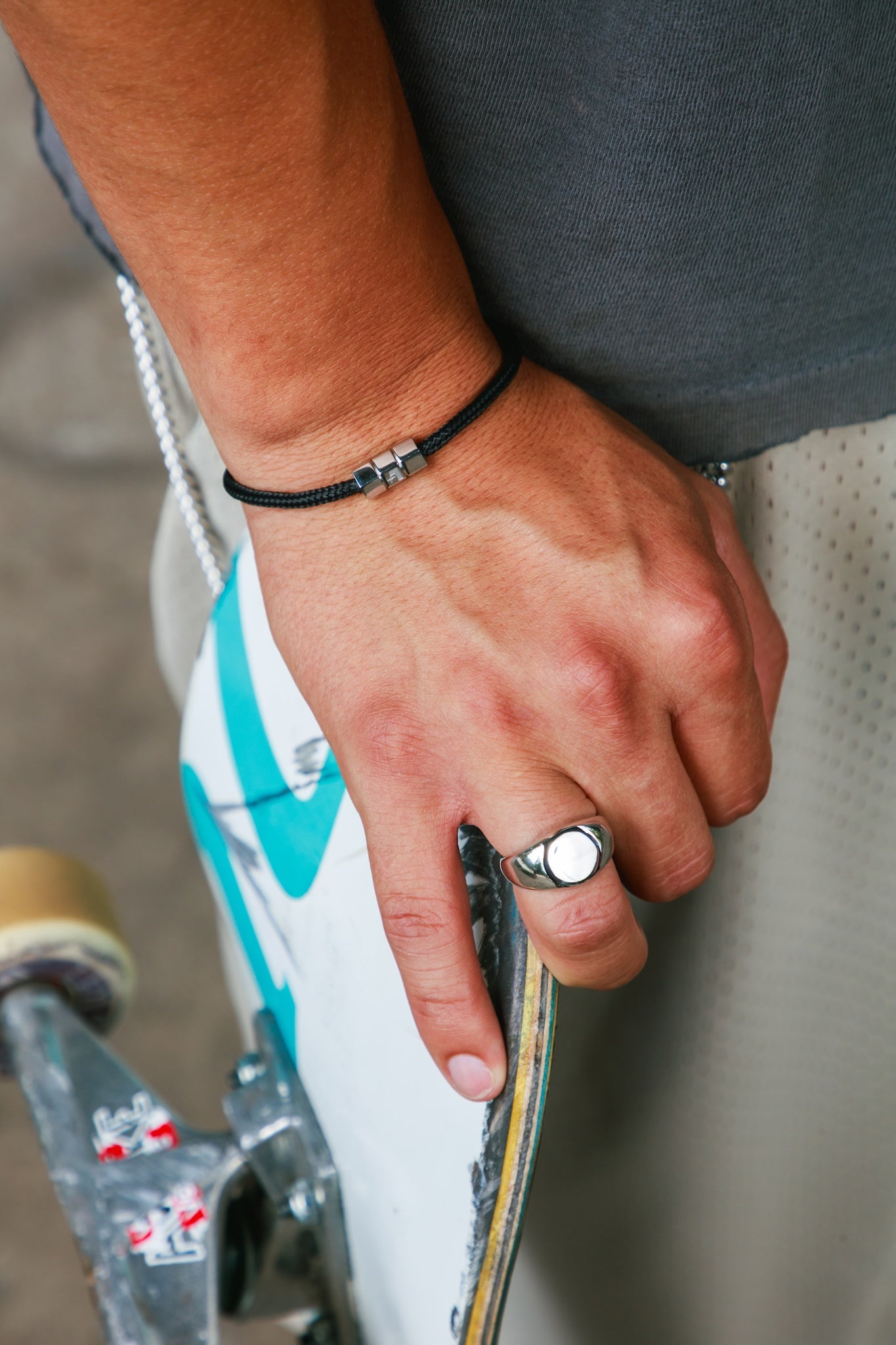 A close-up lifestyle shot of a person's hand resting on a white and teal skateboard, wearing a silver signet ring and a black cord bracelet.