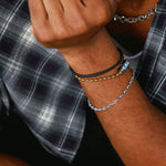 Close-up of a person wearing multiple bracelets on one wrist, including a silver chain bracelet, a black cord bracelet with a gold bead, and a black cord bracelet with a silver bead, paired with a plaid shirt