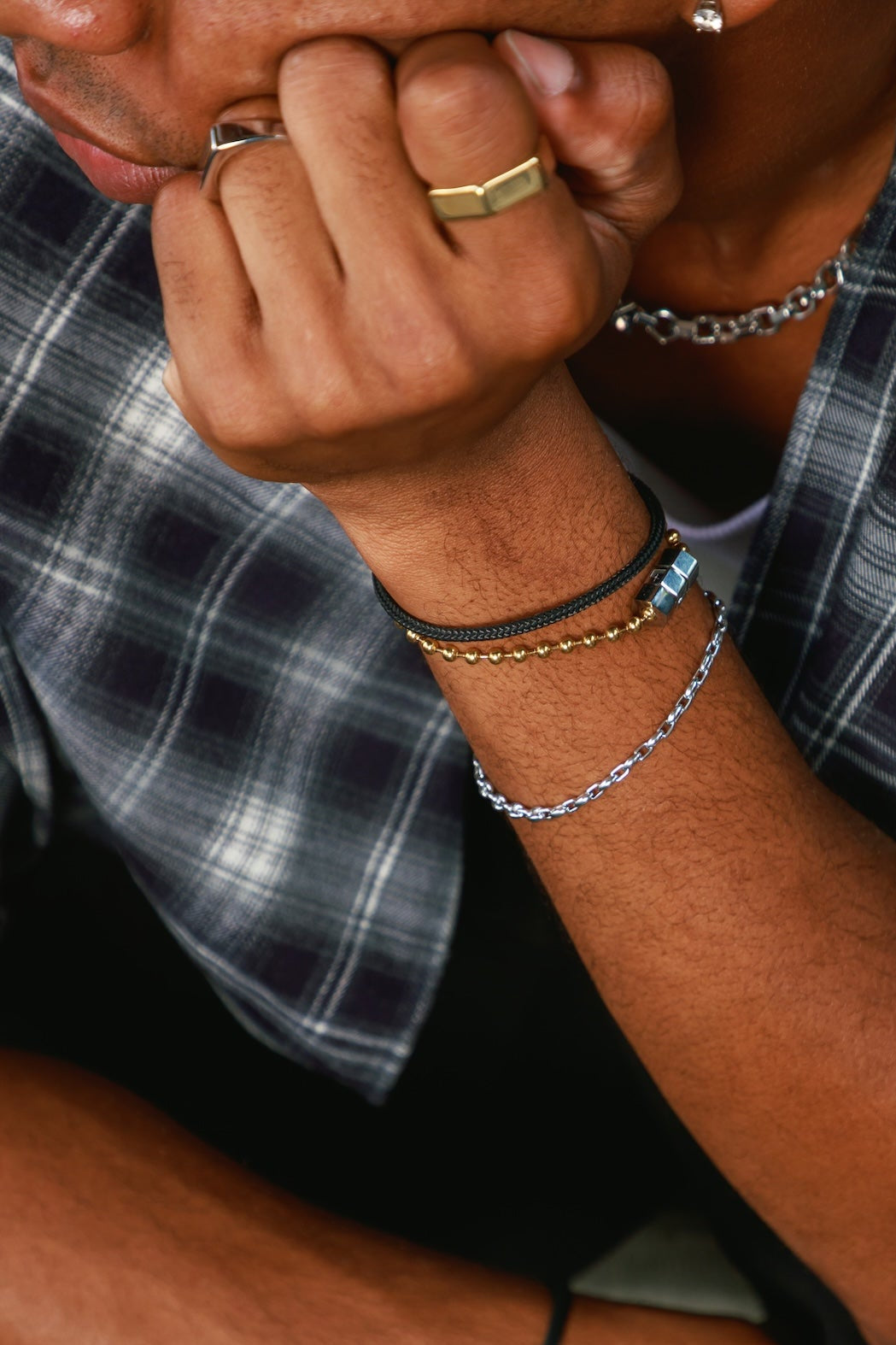 Close-up of a person wearing multiple bracelets on one wrist, including a silver chain bracelet, a black cord bracelet with a gold bead, and a black cord bracelet with a silver bead, paired with a plaid shirt