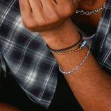 Close-up of a person wearing multiple bracelets on one wrist, including a silver chain bracelet, a black cord bracelet with a gold bead, and a black cord bracelet with a silver bead, paired with a plaid shirt