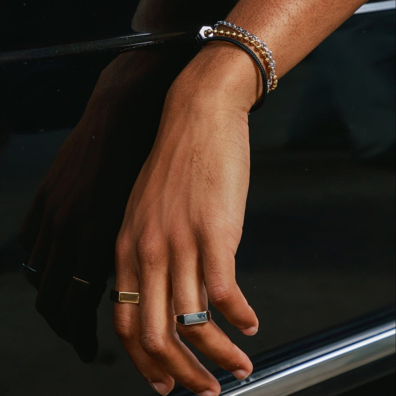 A close-up of a person's hand resting on a glossy black car door, wearing a stack of bracelets on one wrist and two rings on the fingers.